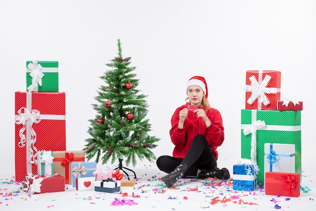 Vista frontale della giovane donna seduta intorno a regali in possesso di carta di credito rossa sul muro bianco