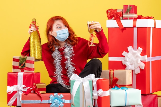 Vista frontale della giovane donna seduta intorno ai regali di Natale in maschera con champagne sulla parete gialla