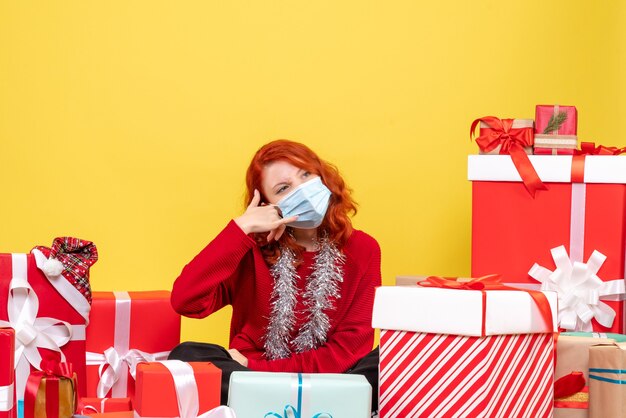 Vista frontale della giovane donna seduta intorno ai regali di Natale in maschera su sfondo giallo nuovo anno virus emozioni covid- colore