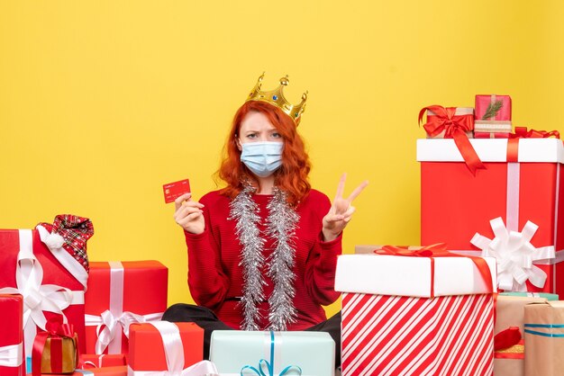 Vista frontale della giovane donna seduta intorno presenta in maschera con carta di credito sulla parete gialla