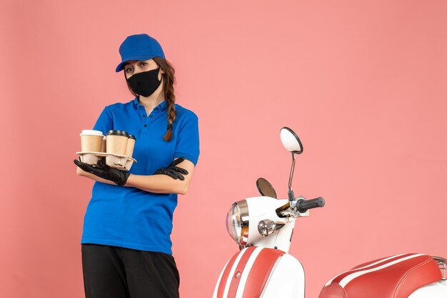 Vista frontale della giovane ragazza del corriere che indossa guanti con maschera medica in piedi accanto alla moto con in mano piccole torte di caffè su sfondo color pesca pastello