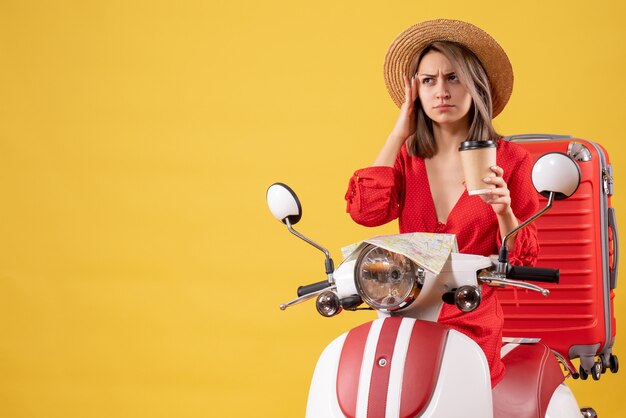 Vista frontale della giovane signora confusa in vestito rosso che tiene la tazza di caffè vicino al ciclomotore