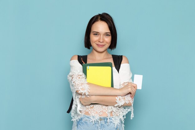 Vista frontale della giovane studentessa in camicia bianca blue jeans e borsa nera che tiene i quaderni e la carta bianca sulla scuola universitaria studentessa dello spazio blu