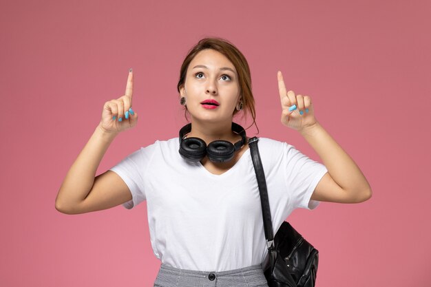 Vista frontale della giovane studentessa in t-shirt bianca e pantaloni grigi con gli auricolari che esamina i cieli su sfondo rosa lezioni di studente University College