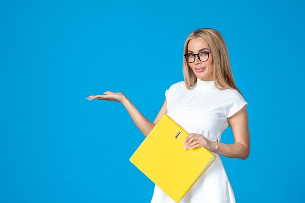 Vista frontale della lavoratrice in abito bianco che tiene cartella gialla sulla parete blu