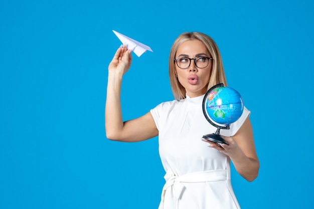 Vista frontale della lavoratrice in abito bianco che tiene il globo terrestre e l'aereo di carta sulla parete blu