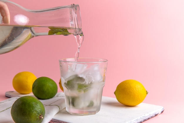 Vista frontale della limonata fredda fresca con ghiaccio all'interno del vetro insieme a limoni freschi sulla parete rosa