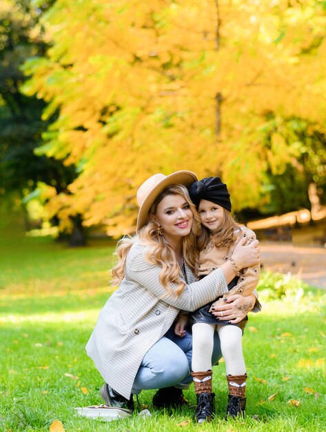 Vista frontale della madre allegra con la figlia adorabile che indossa abbigliamento casual e cappello che si abbracciano e si siedono insieme tra il prato sullo sfondo