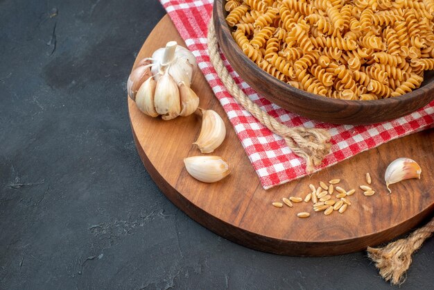 Vista frontale della pasta cruda in una ciotola marrone sul riso rosso dell'aglio dell'asciugamano spogliato sulla corda rotonda del bordo di legno