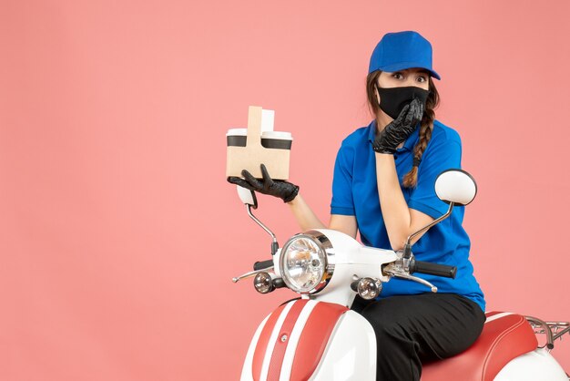 Vista frontale della persona di consegna femminile sorpresa che indossa maschera medica e guanti seduti su uno scooter che consegna ordini su sfondo color pesca pastello