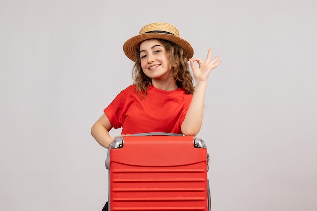 Vista frontale della ragazza di vacanza con la sua valigia facendo segno okey in piedi sul muro bianco