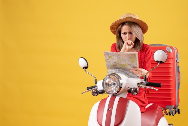 Vista frontale della ragazza graziosa confusa sul ciclomotore con la mappa rossa della tenuta della valigia