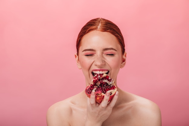 Vista frontale della ragazza nuda che mangia granato. Studio shot di donna con melograno isolato su sfondo rosa.