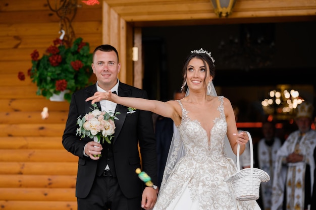 Vista frontale della sposa allegra in abito da sposa che esce dall'edificio tenendo il cesto e lanciando dolci agli ospiti mentre il suo sposo in piedi dietro e tiene in mano il bouquet da sposa dopo la cerimonia del matrimonio