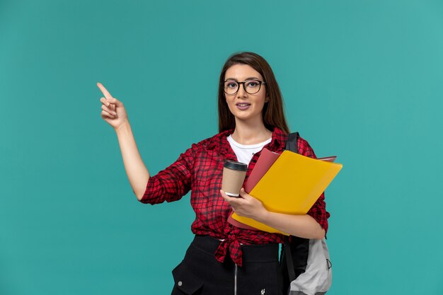 Vista frontale della studentessa che indossa uno zaino che tiene file e caffè sulla parete blu