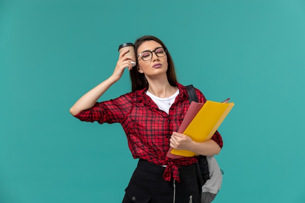 Vista frontale della studentessa che indossa uno zaino che tiene file e caffè sulla parete blu