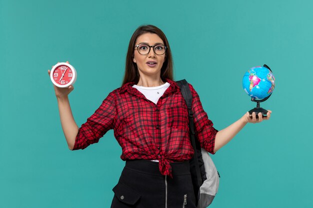 Vista frontale della studentessa che indossa uno zaino che tiene piccolo globo e orologi sulla parete blu chiaro