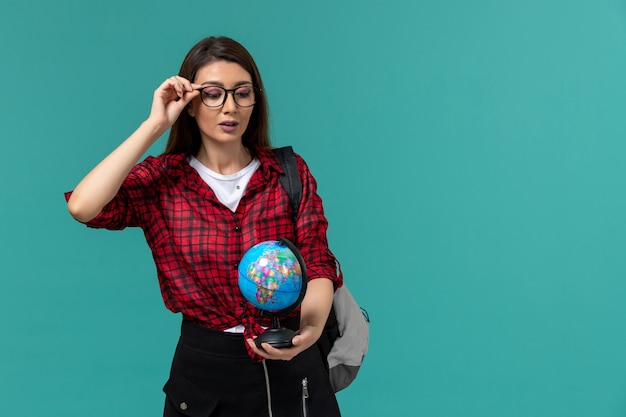 Vista frontale della studentessa che indossa uno zaino che tiene piccolo globo sulla parete blu-chiaro