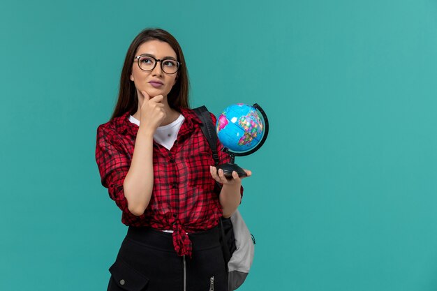 Vista frontale della studentessa che indossa uno zaino che tiene piccolo globo sulla parete blu-chiaro