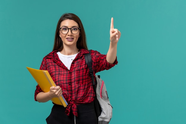 Vista frontale della studentessa che indossa uno zaino e che tiene i file sorridente sulla parete azzurra