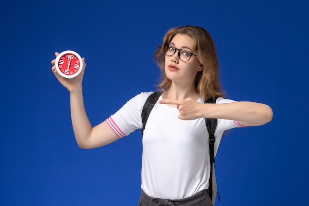 Vista frontale della studentessa in camicia bianca che indossa uno zaino e tenendo gli orologi sulla scrivania blu