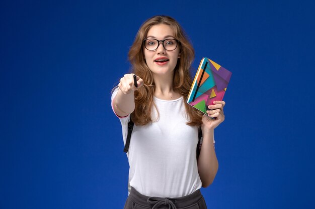 Vista frontale della studentessa in camicia bianca che tiene penna e quaderno sulla parete blu
