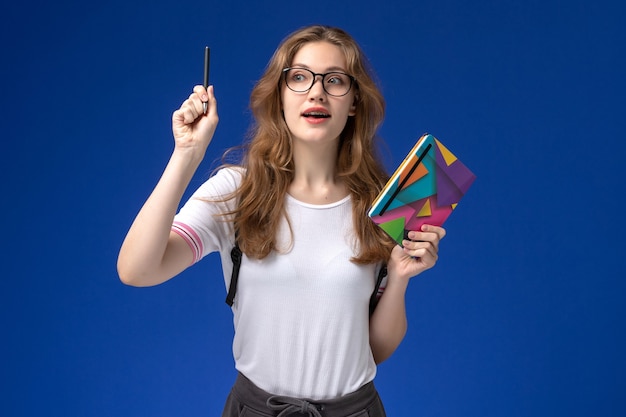 Vista frontale della studentessa in camicia bianca che tiene penna e quaderno sulla parete blu