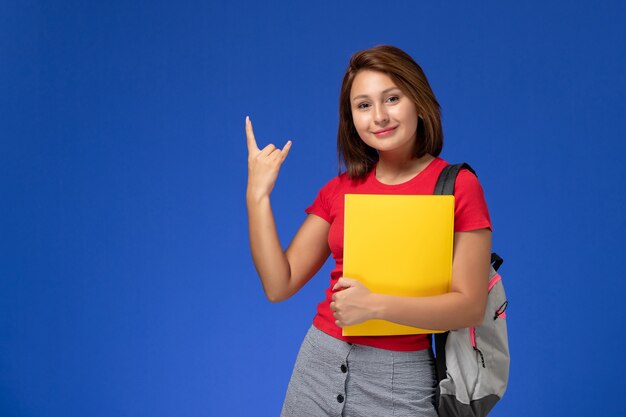 Vista frontale della studentessa in camicia rossa con lo zaino che tiene file gialli sulla parete azzurra