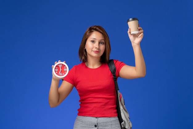 Vista frontale della studentessa in camicia rossa con lo zaino che tiene gli orologi e il caffè sulla parete blu