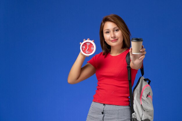 Vista frontale della studentessa in camicia rossa con lo zaino che tiene gli orologi e il caffè sulla parete blu