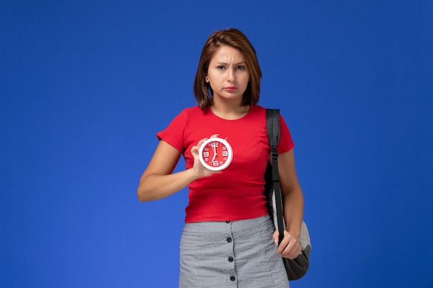 Vista frontale della studentessa in camicia rossa con lo zaino che tiene gli orologi sulla parete blu