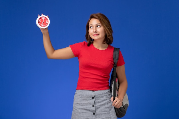 Vista frontale della studentessa in camicia rossa con lo zaino che tiene gli orologi sulla parete blu