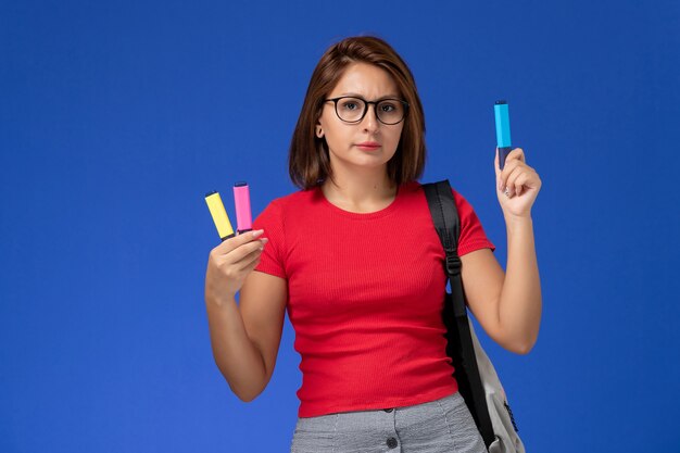 Vista frontale della studentessa in camicia rossa con lo zaino che tiene i pennarelli sulla parete blu