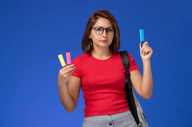 Vista frontale della studentessa in camicia rossa con lo zaino che tiene i pennarelli sulla parete blu