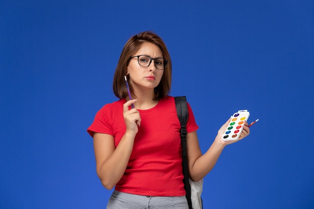 Vista frontale della studentessa in camicia rossa con lo zaino che tiene le vernici per il disegno e le nappe sulla parete blu