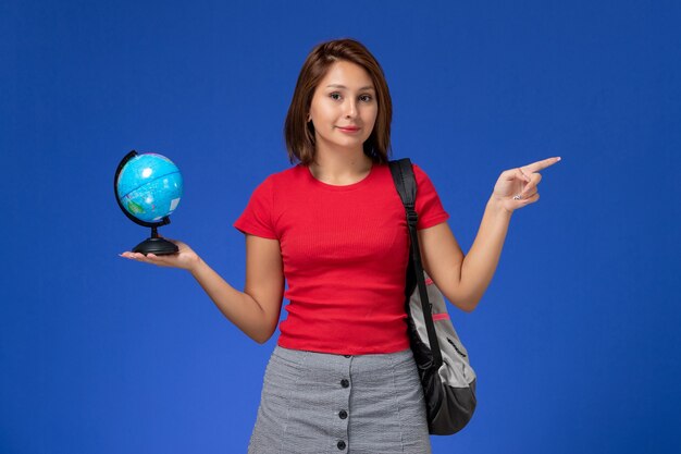 Vista frontale della studentessa in camicia rossa con lo zaino che tiene piccolo globo sulla parete blu-chiaro