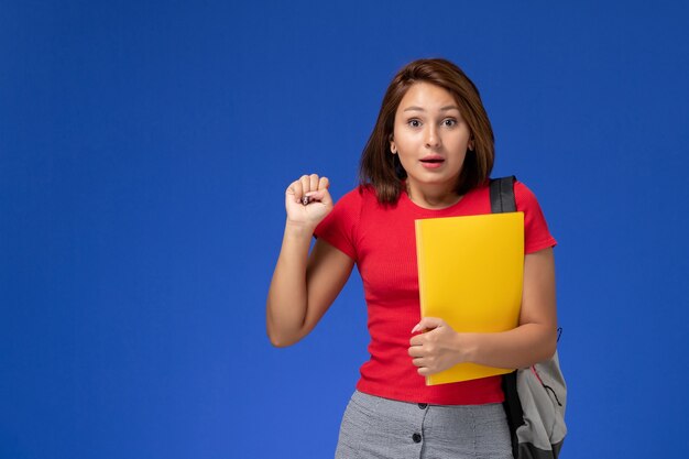 Vista frontale della studentessa in camicia rossa con zaino che tiene file gialli sulla parete blu