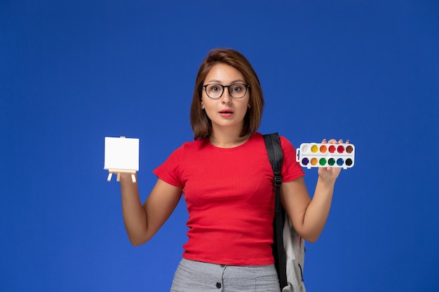 Vista frontale della studentessa in camicia rossa con zaino che tiene le vernici per disegnare sulla parete blu