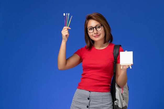 Vista frontale della studentessa in camicia rossa con zaino che tiene nappe per disegnare sorridente sulla parete azzurra