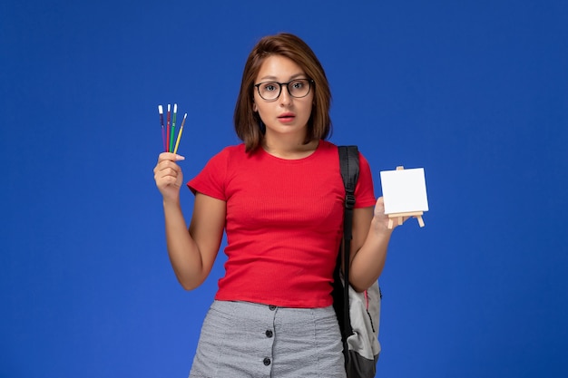 Vista frontale della studentessa in camicia rossa con zaino che tiene nappe per disegnare sulla parete azzurra