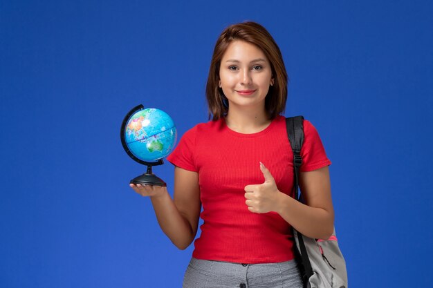 Vista frontale della studentessa in camicia rossa con zaino che tiene piccolo globo sorridente sulla parete blu