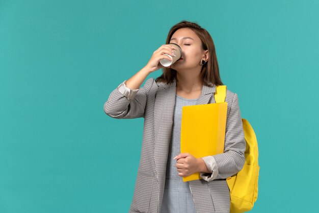 Vista frontale della studentessa in giacca grigia che indossa il suo zaino giallo con file di bere il caffè sulla parete azzurra
