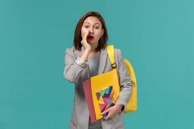 Vista frontale della studentessa in giacca grigia che indossa lo zaino giallo che tiene i file e il quaderno che bisbiglia sulla parete blu