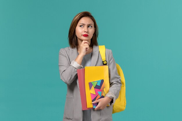 Vista frontale della studentessa in giacca grigia che indossa uno zaino giallo che tiene i file e il quaderno pensando sulla parete blu