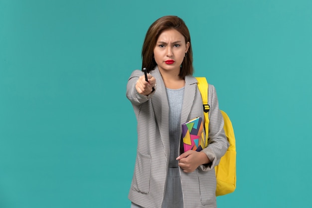 Vista frontale della studentessa in giacca grigia che indossa uno zaino giallo che tiene il quaderno con la penna sulla superficie blu