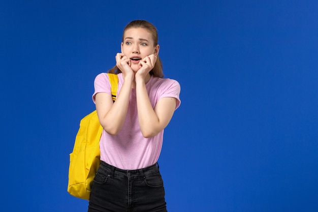 Vista frontale della studentessa in maglietta rosa con zaino giallo con espressione spaventata sulla parete blu