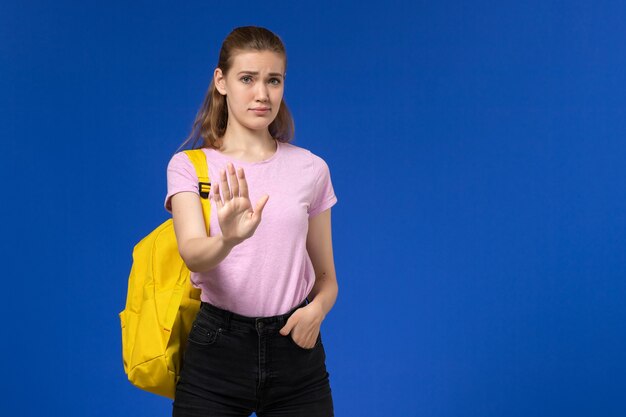 Vista frontale della studentessa in maglietta rosa con zaino giallo in posa sulla parete azzurra