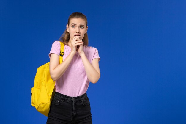 Vista frontale della studentessa in maglietta rosa con zaino giallo in posa sulla parete blu