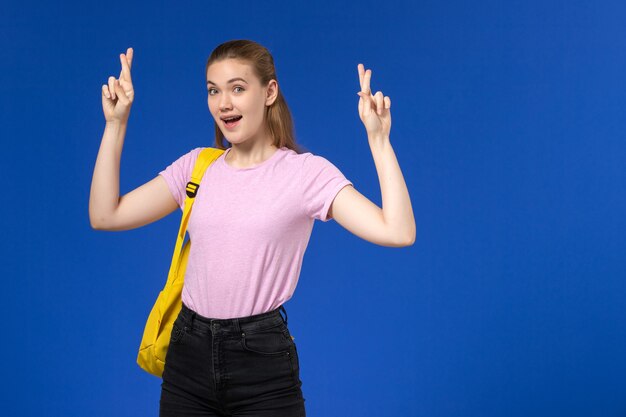 Vista frontale della studentessa in maglietta rosa con zaino giallo incrociando le dita sulla parete blu