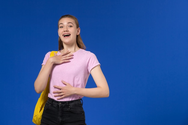 Vista frontale della studentessa in maglietta rosa con zaino giallo sulla parete blu